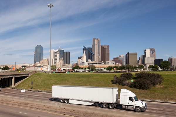 A white semi-trailer truck driving on a multi-lane highway with the downtown Dallas, Texas skyline visible in the background.