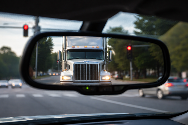 A view through a car's rearview mirror showing a large white semi-truck tailgating closely behind on a Fort Worth, TX, city street, with red traffic lights and other cars visible in the background.